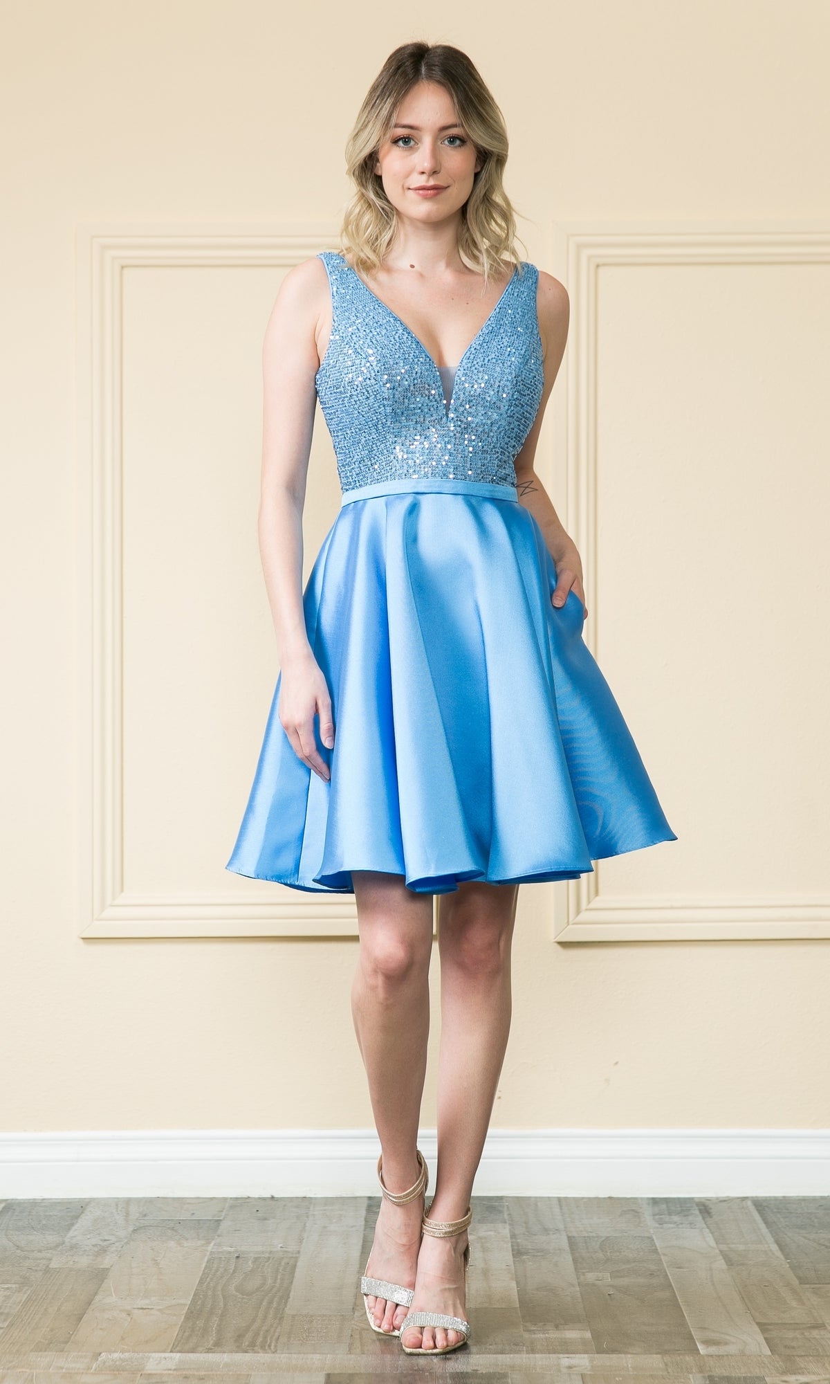 A woman stands indoors wearing the A-Line Short Homecoming Dress with Pockets - 8954, featuring a sequined, embellished bodice and flared skirt, paired with strappy heels.